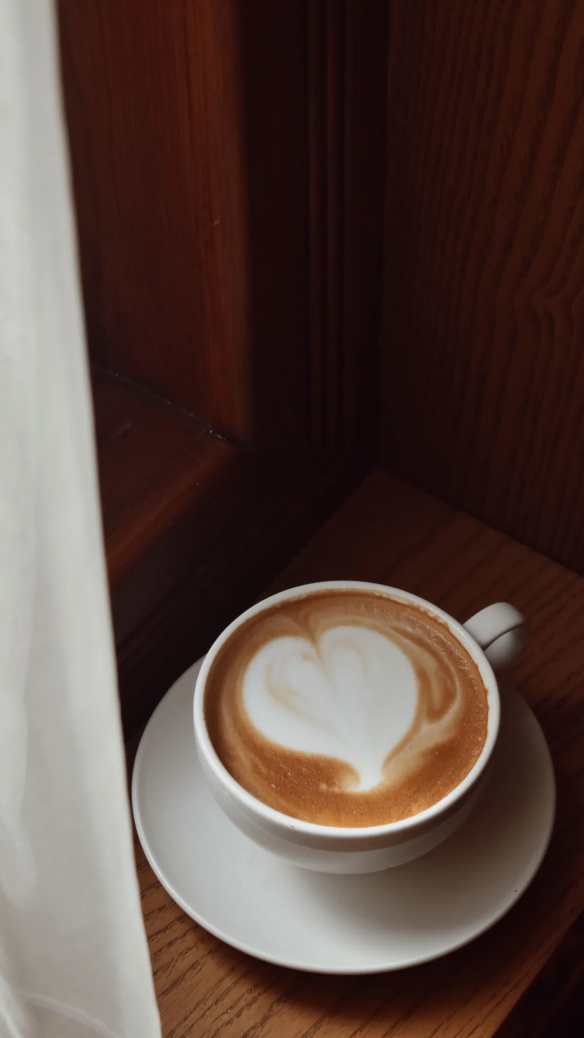 Cappuccino com desenho de coração em mesa próxima à janela